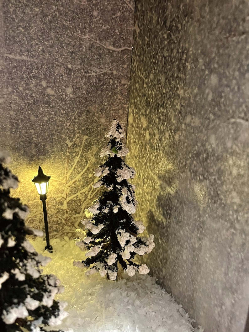 Op de afbeelding: Een besneeuwde wintersc&egrave;ne met twee kleine, met sneeuw bedekte dennenbomen en een verlichte lantaarnpaal. De achtergrond is een getextureerde, grijze muur.