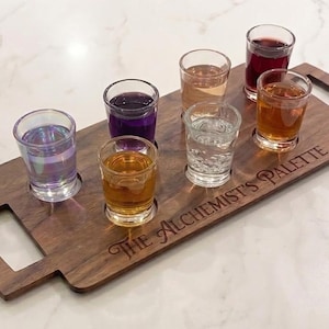 Puede incluir: Una bandeja de madera con siete vasos de chupito llenos de líquidos de colores. La bandeja tiene un asa y el texto "The Alchemist's Palette" grabado. Los vasos son transparentes y tienen una base gruesa.