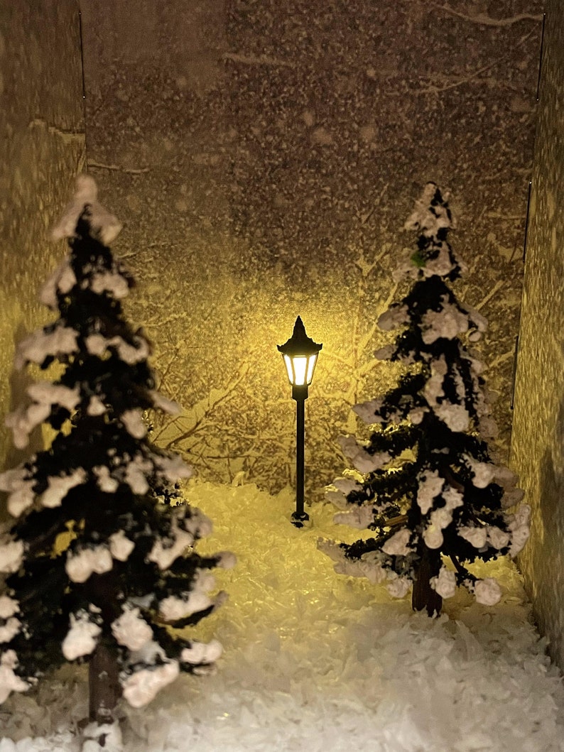 Op de afbeelding: Een miniatuur wintersc&egrave;ne met twee met sneeuw bedekte dennenbomen en een verlichte lantaarnpaal in het midden. De sc&egrave;ne is geplaatst op een wit, pluche oppervlak dat op sneeuw lijkt.