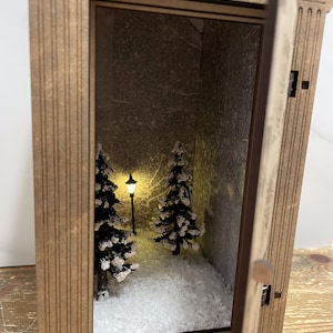Op de afbeelding: Een houten lantaarn met een sneeuwsc&egrave;ne erin. De lantaarn heeft een klein deurtje dat open staat, waardoor een miniatuur winterlandschap zichtbaar wordt met twee dennenbomen en een brandende lantaarnpaal. De sc&egrave;ne is bedekt met witte glitter om sneeuw na te bootsen.
