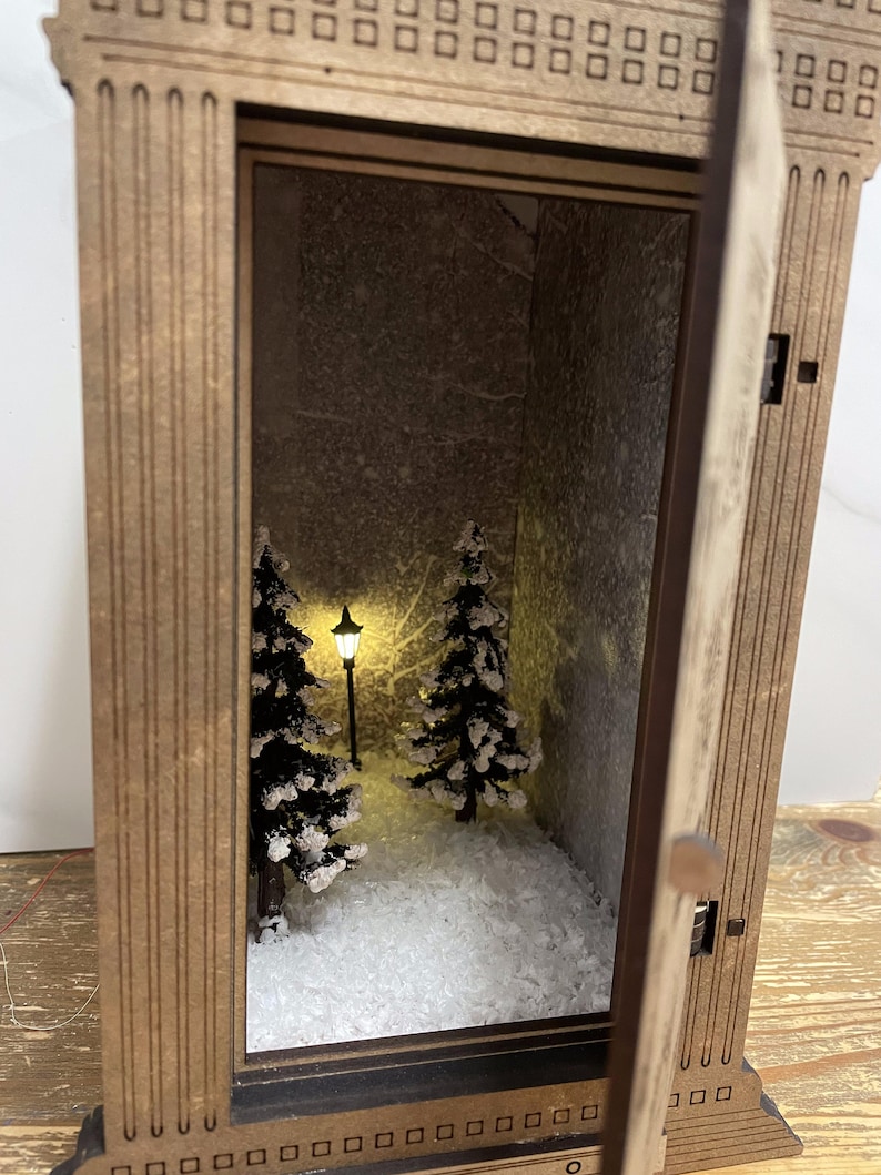 Op de afbeelding: Een houten lantaarn met een sneeuwsc&egrave;ne erin. De lantaarn heeft een klein deurtje dat open staat, waardoor een miniatuur winterlandschap zichtbaar wordt met twee dennenbomen en een brandende lantaarnpaal. De sc&egrave;ne is bedekt met witte glitter om sneeuw na te bootsen.
