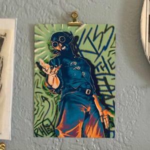 Pode incluir: Uma impress&atilde;o art&iacute;stica retangular apresentando uma figura usando uma m&aacute;scara de g&aacute;s escura e um uniforme correspondente com cinto. A figura posa com as m&atilde;os estendidas contra um fundo verde claro com texto azul estilo grafite KISS THE PAIN.