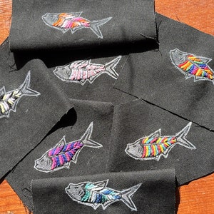 May include: Black fabric patches embroidered with colorful fish designs. Each fish is outlined in white and filled with various colors, including pink, blue, yellow, and green. The patches are arranged on a wooden surface.