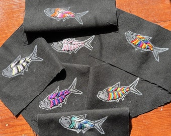 Pode incluir: Remendos de tecido preto bordados com desenhos coloridos de peixes. Cada peixe é delineado em branco e preenchido com várias cores, incluindo rosa, azul, amarelo e verde. Os remendos estão dispostos sobre uma superfície de madeira.