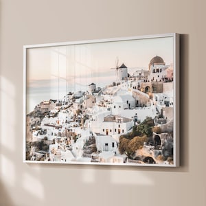 May include: A framed print depicting a coastal village with white buildings on a hillside. The image shows windmills, a domed structure, and a warm colour palette. The print is in a white frame.