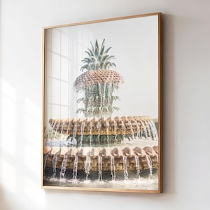 May include: Framed print of a pineapple fountain with cascading water. The fountain has a detailed, textured design and is set against a bright, neutral background. The frame is a light wood colour.