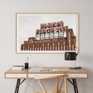 以下が含まれることがあります： レンガ造りの建物にある「Farine Five Roses」の看板の額入りプリント。看板は赤と白です。プリントは、黒いランプと籐の椅子を備えた木製の机の上に掛けられています。