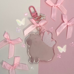 May include: Pink and white capybara charm with wings, attached to a pink ribbon and a silver keyring. The charm is surrounded by pink bows and glitter.