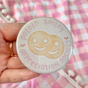 Peut inclure: Un badge rond de couleur claire avec l'inscription "potato smiley appreciation society" en rose. Deux visages de pommes de terre souriants sont au centre. Le fond est un motif de grille claire.