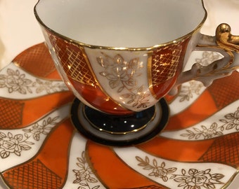 May include: A white teacup and saucer set with an orange and white geometric pattern and gold accents. The cup has a gold-trimmed handle and a black base.