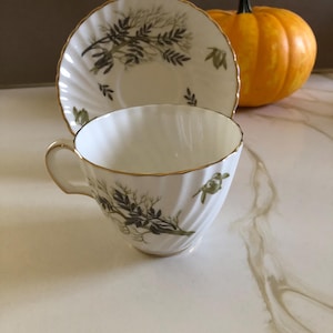 May include: A white teacup and saucer set with a gold rim and a green and black botanical design. The teacup has a curved handle and a fluted body. A small orange pumpkin is in the background.