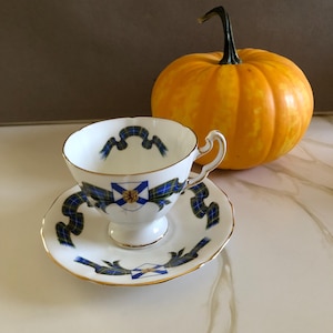 May include: A white teacup and saucer set with a gold trim and a blue and green plaid ribbon design. A small orange pumpkin sits behind the teacup. The teacup is on a white surface.