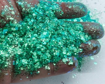 May include: A close-up of a hand holding a pile of iridescent green glitter. The glitter is a mix of different sizes and shapes, and it has a shimmery finish.