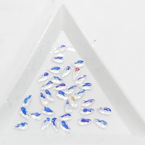 May include: A collection of iridescent teardrop-shaped rhinestones in various sizes, scattered on a white surface.