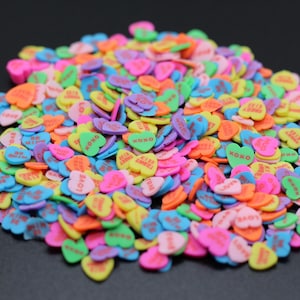May include: A large pile of colorful heart-shaped sprinkles with various messages like "XOXO", "Love", and "Be Mine".