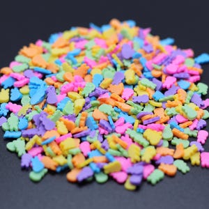 May include: A large pile of colorful sprinkles in various shapes, including hearts, stars, and other abstract shapes. The sprinkles are a mix of pink, blue, green, orange, and yellow.