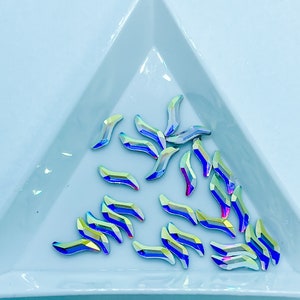 May include: A collection of iridescent blue and green crystal rhinestones in a teardrop shape with a pointed tip. The rhinestones are arranged in a random pattern on a light blue surface.