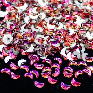 May include: A large pile of iridescent crescent-shaped rhinestones in shades of pink and orange. The rhinestones are scattered on a black surface.