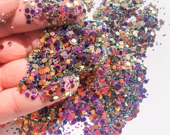 May include: A close-up of iridescent glitter in shades of purple, gold, and orange. The glitter is scattered on a white surface and is being held in a hand.