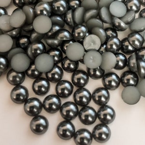 May include: A large pile of gray pearl beads, some with a flat back, and some with a rounded back. The beads are scattered on a white surface.