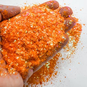 May include: Orange and iridescent glitter in a variety of sizes. The glitter is being held in a hand.