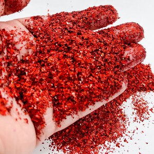 May include: A close-up of a hand holding a pile of red glitter. The glitter is iridescent and has a chunky texture.