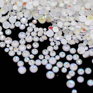 May include: A large quantity of iridescent white round rhinestones, some with a pearlescent finish, scattered on a black surface.
