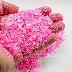 May include: A close-up of a hand holding a large amount of pink glitter with iridescent accents. The glitter is scattered on a white surface.