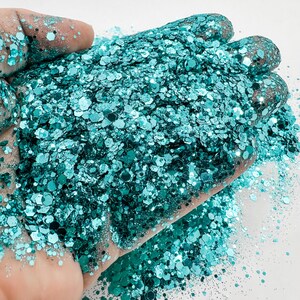 May include: A close-up of teal glitter with a holographic finish. The glitter is in a variety of sizes and shapes.