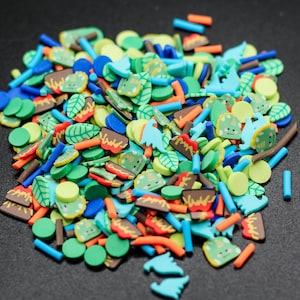 May include: A colorful assortment of polymer clay slices in various shapes and colors. The mix includes green leaves, blue cylinders, orange carrots, brown tree trunks, and light green round shapes. Some slices have cartoon faces.