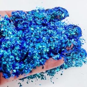 May include: A close-up of a hand holding a pile of blue and teal iridescent glitter. The glitter is made up of small, irregular pieces that catch the light and sparkle.