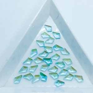 May include: A pile of light blue iridescent diamond-shaped rhinestones in a white triangular tray.