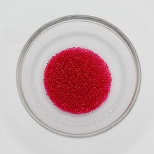 May include: A clear glass bowl filled with a large cluster of small, bright red beads. The beads are densely packed, forming a circular shape within the bowl. The bowl sits on a white surface.