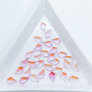 May include: A collection of iridescent pink and orange diamond-shaped rhinestones in a white triangular tray.