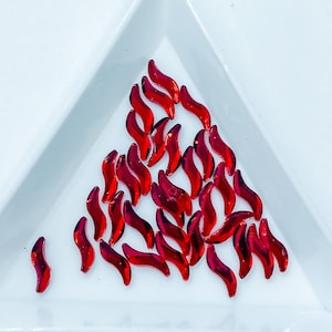 May include: A collection of red, iridescent, teardrop-shaped rhinestones on a white surface.
