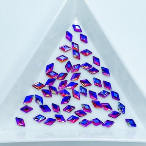 May include: A pile of iridescent purple and pink diamond-shaped rhinestones on a white triangular surface.