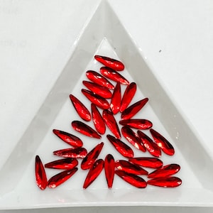 May include: A collection of red, teardrop-shaped glass rhinestones, scattered on a white triangular surface.
