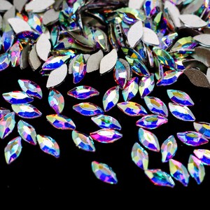 May include: A scattering of iridescent, teardrop-shaped rhinestones on a black background. The rhinestones have a rainbow-like sheen.