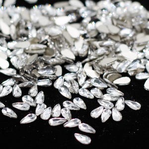 May include: A collection of clear, teardrop-shaped rhinestones with a flat, silver backing. The rhinestones are scattered across a black surface, reflecting light and creating a sparkling effect. Ideal for embellishing crafts or adding sparkle to garments.