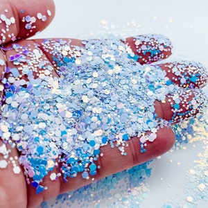 Puede incluir: Primer plano de una mano sosteniendo un mont&oacute;n de purpurina azul y blanca iridiscente. La purpurina es una mezcla de hex&aacute;gonos y c&iacute;rculos peque&ntilde;os y grandes.