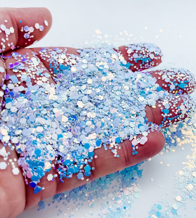 Puede incluir: Primer plano de una mano sosteniendo un mont&oacute;n de purpurina azul y blanca iridiscente. La purpurina es una mezcla de hex&aacute;gonos y c&iacute;rculos peque&ntilde;os y grandes.