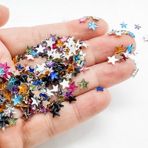 May include: A handful of colorful star-shaped sequins in various colors, including pink, blue, silver, and gold. The sequins are scattered across a light background, creating a sparkling effect. These decorative embellishments are ideal for crafts and DIY projects.