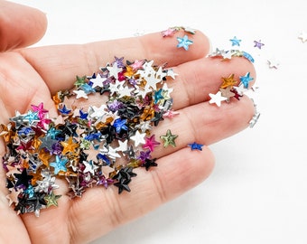 May include: A handful of colorful star-shaped sequins in various colors, including pink, blue, silver, and gold. The sequins are scattered across a light background, creating a sparkling effect. These decorative embellishments are ideal for crafts and DIY projects.