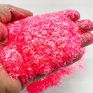 May include: A handful of vibrant pink glitter, composed of small, elongated pieces. The glitter has a bright, neon color and a reflective quality, catching the light. The image is a close-up, highlighting the texture and color of the glitter.