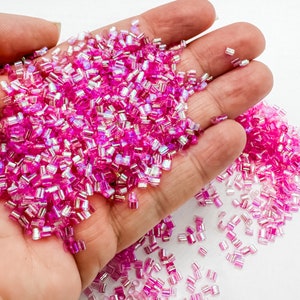 May include: A handful of vibrant pink, iridescent seed beads. The small, rectangular beads are scattered across a hand, with some spilling out. The beads have a shiny, reflective quality, catching the light and creating a shimmering effect. Ideal for jewelry making and crafts.