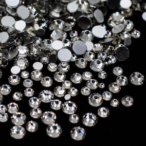 May include: A close-up of a large quantity of clear crystal rhinestones of various sizes scattered on a black surface.