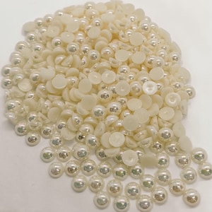 May include: A pile of ivory-colored half-round and round faux pearls. The pearls have a shiny, iridescent finish, reflecting light. The flat-backed half-round pearls are mixed with the round pearls.