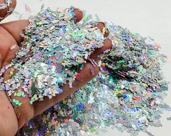 May include: A large amount of iridescent silver glitter in the shape of candy. The glitter is scattered on a white surface and is being held in a hand.
