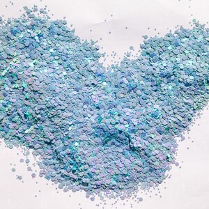 May include: A close-up of iridescent blue glitter, scattered on a white surface. The glitter is in the shape of a heart.