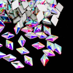 May include: A scattering of iridescent, faceted, diamond-shaped rhinestones on a black background. The rhinestones are various shades of pink, purple, and blue.